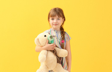 Cute little girl holding nebulizer and toy bear on color background. Allergy concept