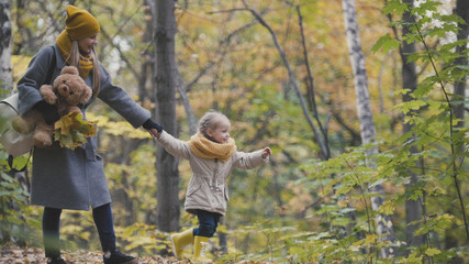 Naklejka na ściany i meble Little daughter with her mommy and Taddy bear walks in autumn park - plays the leaves