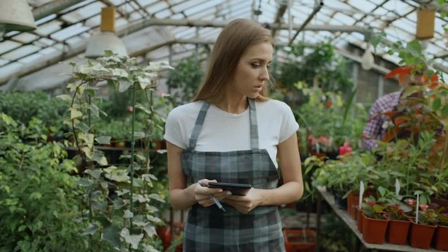 Dolly shot of Young woman working in garden center. Attractive girl check and count flowers using tablet computer during work in greenhouse