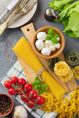 Raw ingredients of Italian pasta, spaghetti, tagliatelle, fusilli, garlic, basil, mozzarella,  pepper, cherry tomatoes and olive oil on gray  background. Selective focus.Top view. Copy space.