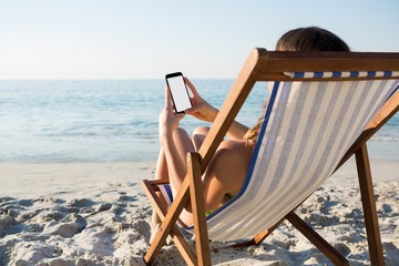 Fototapeta premium Kobieta używa telefon komórkowego podczas gdy relaksujący na holu krześle przy plażą