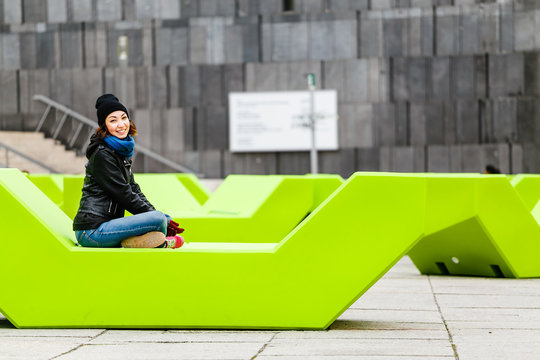 Teenage girl in casual clothes sitting on vivid green unusual bench at city street