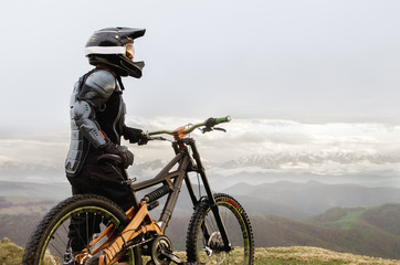 Fototapeta premium The rider in the full-face helmet and full protective equipment on the mtb bike stands on a rock against the background of a ridge and low clouds