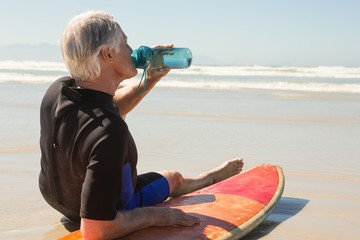senior man drinking water while sitting on surfboard