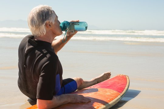 senior man drinking water while sitting on surfboard