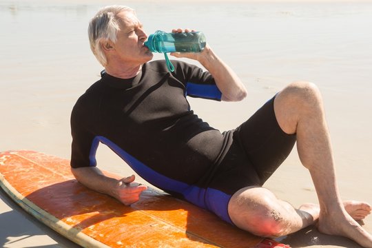 Senior man drinking water while sitting by surfboard
