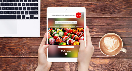 Person using modern tablet for searching food online, placed on wooden table with laptop and cup of coffee