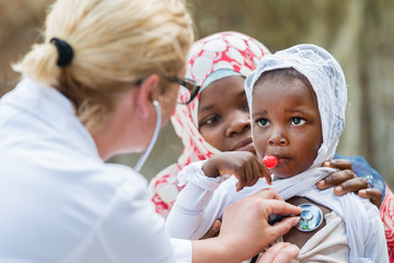 Female Caucasian doctor listening heart beat and breathing of little African girl with...