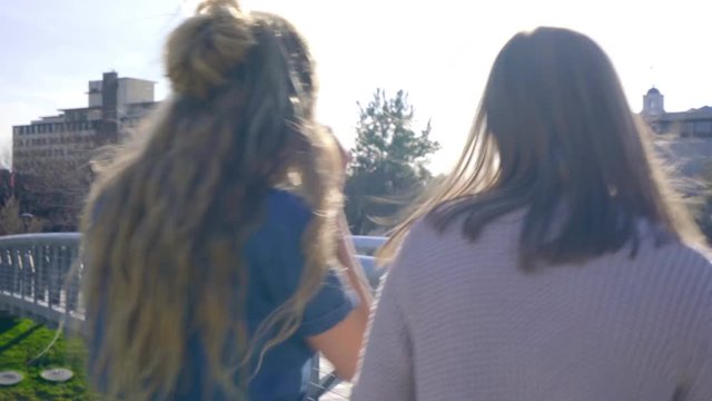 Teen Girls Share Headphones, Listen To Music On Smartphone, Walk Across Bridge In City Park