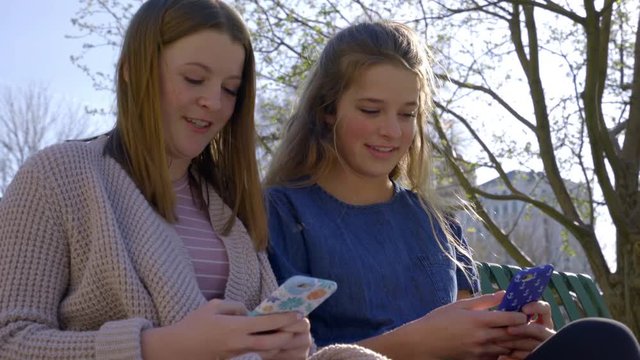 Two Teen Girls Sit On City Park Bench And Enjoy Using Smartphones, They Look At Photos And Laugh