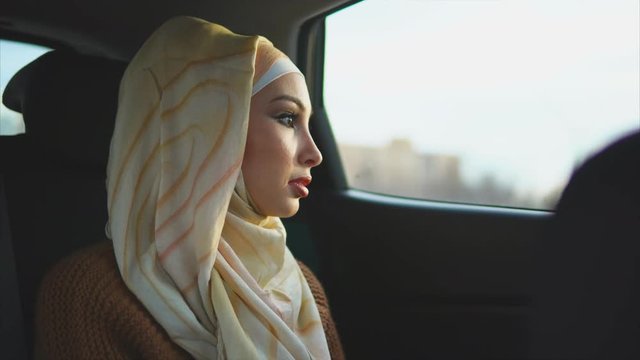 Young Muslim woman traveling on backseat of a taxi. She enjoying city view through the car window and laughing at the end
