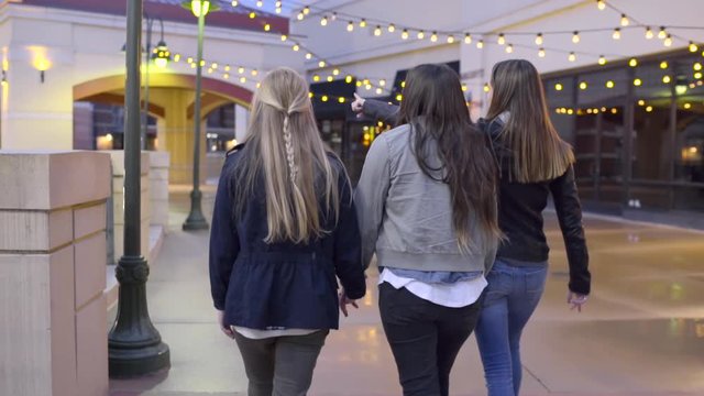 Teens Walk Around Outdoor Mall At Night, Girls Hold Hands, Their Friend Points To Something In Distance
