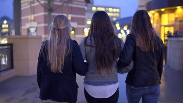 Slow Motion Shot Of Teen Girls Walking Arm In Arm (Away From Camera) Past Shops In City 