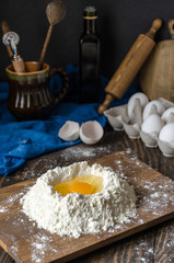 Egg yolk in flour close up on a wooden table. In the background rolling pin, board, olive oil, spoon and ravioli knife and eggs