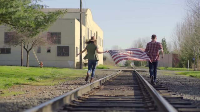 Couple Run Away From Camera, Down Train Tracks, Holding American Flag Between Them