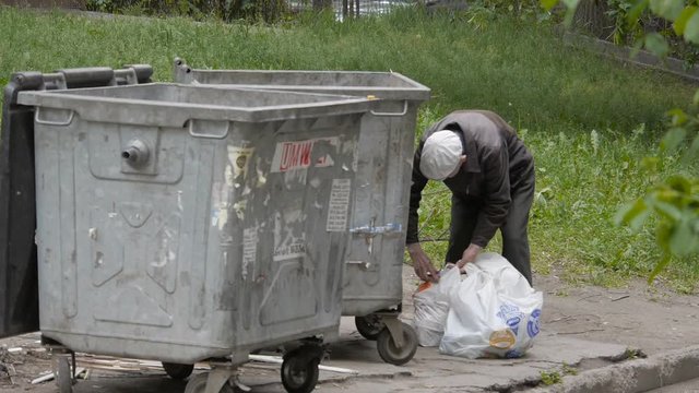 Homeless, infirm and old man puts in packages а waste, waste paper near garbage cans
