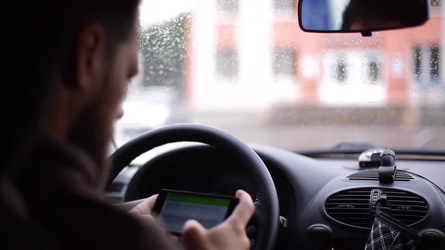 A man with a beard sits in a car and writes an SMS message on a smartphone