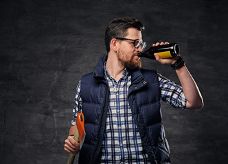A man dressed in fleece shirt holds an axe and drinks craft bottled beer.