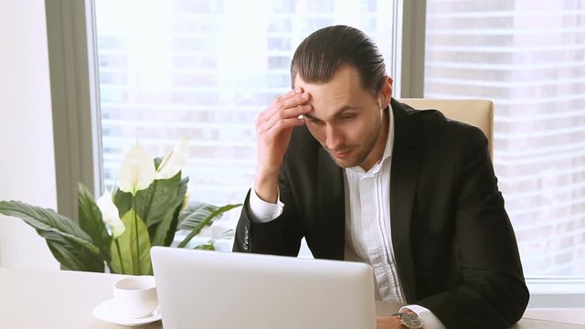 Overworked young businessman yawning, looking at wristwatch and checking time at office using laptop, feeling tired exhausted, working extra hours, unmotivated employee, suffering from lack of sleep