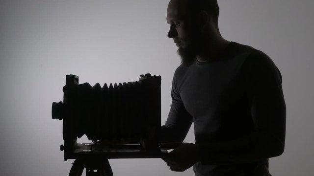 Mid shot of bearded photographer adjusting old vintage wooden photo camera and taking picture against the light in the black background.