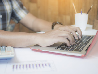 Close up of Asian hipster business man typing on laptop keyboard for working, woking male using notebook to analyze on a desk with a wooden wall