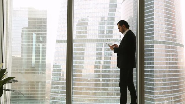 Confident young businessman wearing suit standing near full length window in office, holding tablet or smartphone, looking at big city urban buildings, texting messages, chatting or using business