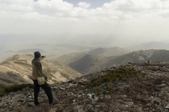 A man stands on top of a mountain and points a finger forward, towards the mountains forming a surprisingly beautiful landscape