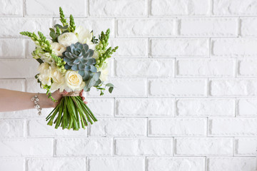 Naklejka premium Female hand gives bunch with roses and succulent on white brick background. Gift for mother or woman, florist work, wedding decor, beautiful bouquet sale concept