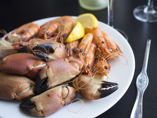 Boiled crab claws, whole prawns and slices of lemon on white plate with a wine glass in the background