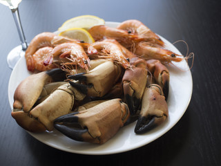 Boiled crab claws, whole prawns and slices of lemon on white plate with a wine glass in the background