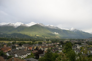 Fototapeta na wymiar Landscape of mountains of a strong spring green, with snow-covered peaks after a strong thunderstorm of mountain and some rays of sunshine, in the foreground valley and houses , alps, italy