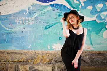 Blonde woman on black dress, necklaces and hat against graffiti wall.