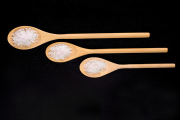 White sea bath salt in wooden spoons on a dark polished table.