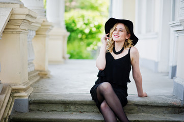 Blonde woman on black dress, necklaces and hat against vintage house.