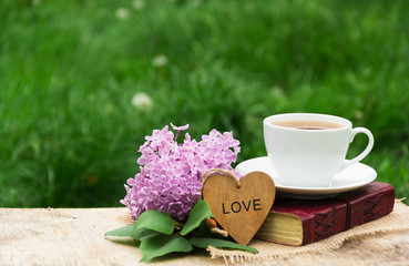 A cup of hot tea, a book and lilacs against a background of green grass. Romantic concept. Wooden card with a heart. Copy space