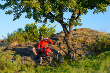 Cyclist in Red Jacket and Helmet Resting and Drinking the Water near Mountain Bike on Rocky Hill...