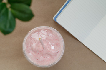 Top view of a strawberry smoothie in a plastic cup with a straw on the table with notebook. Summer & healthy lifestyle concept.