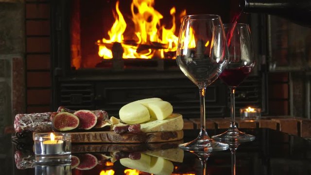 Red wine is poured into glasses on the background of snacks around fireplace