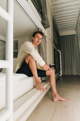 Man sitting in a stylish hostel bedroom. Young man reading book in hotel room with two-levels beds. Evening, grey curtain and wall