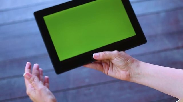 Young white woman holds tablet pc in hands isolated at wooden background. Girl browses internet or watches video. Blank green screen. Point of view video of adult hands of woman and modern touchpad.