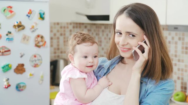 A beautiful and young mother is holding her little baby in her arms while talking on the phone. Modern mom holds a daughter in her arms and uses a mobile phone. Young woman with baby and phone.