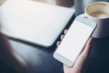 Mockup image of business woman holding mobile phone with blank white screen with latop and coffee cup on wooden table in cafe