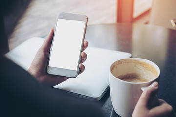 Mockup image of business woman holding mobile phone with blank white screen with latop and coffee cup on wooden table in cafe