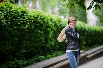 Stylish blonde woman wear at jeans, glasses and leather jacket with phone at hand, against bushes at street. Fashion urban model portrait.