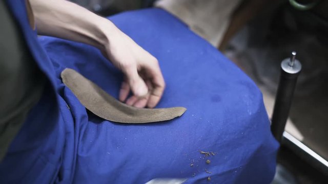 Close up of shoemaker s hands levelling a piece of leather on his lap with a knife. He is wearing a blue apron. Handheld real time close up shot