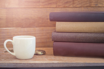stack of thick books with white cup and pocket watch on vintage wooden wall background