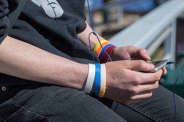 Teenager holding smart phone in hands and listening to music. Both wrists decorated with colorful synthetic leather wristbands. Earphones connected to the gadget with broken glass surface