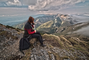 Naklejka na ściany i meble Rear view of a woman sitting on a mountain top peacefully gazing at low-lying clouds. HDR image.