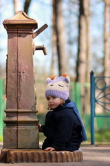 Little girl in coat and hat sitting near the water well