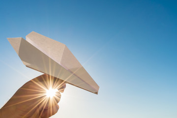 Hand holds white paper plane on blue sky and sun